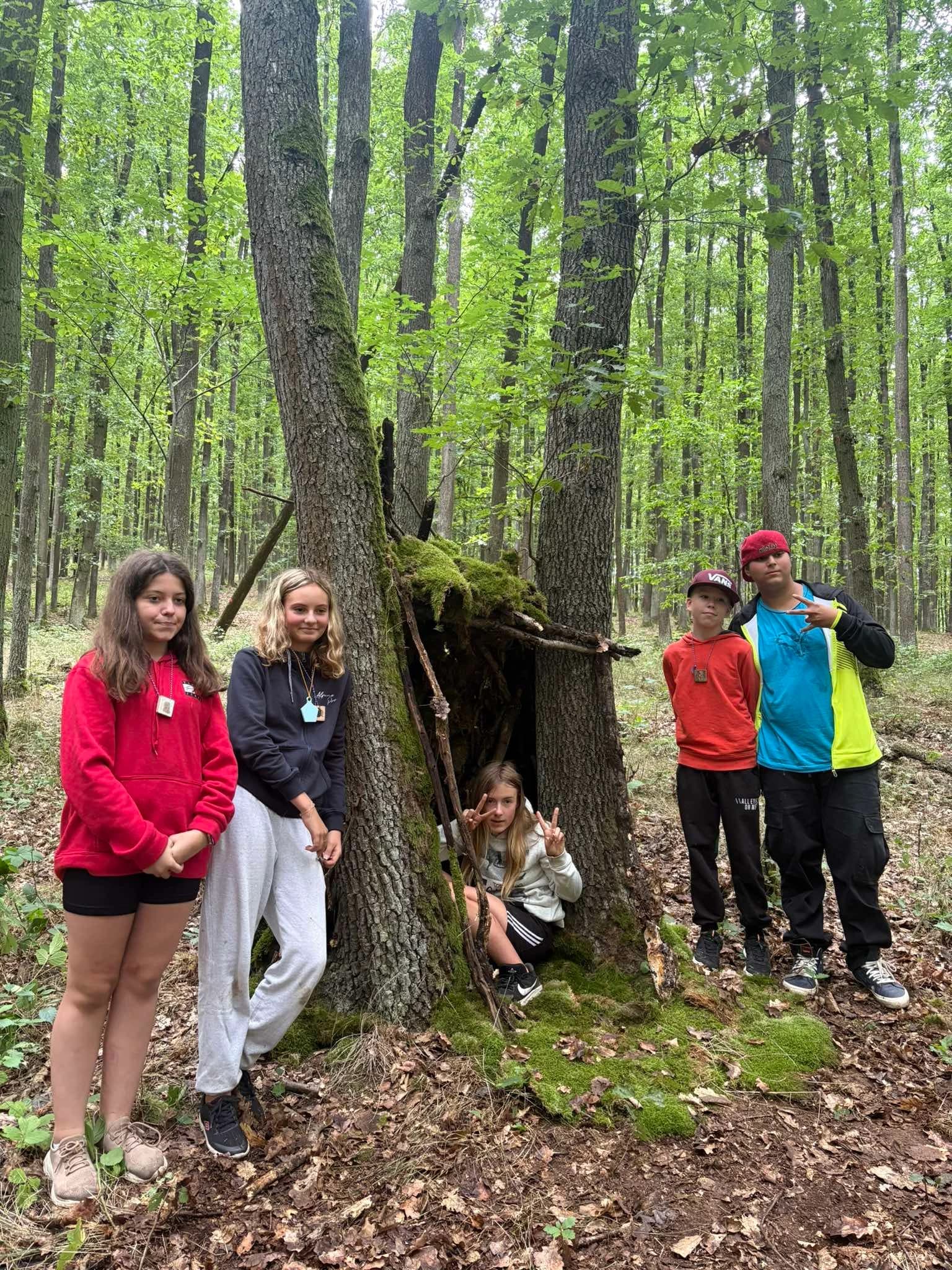 Skupina dětí u postaveného přístřešku mezi stromy 🏕